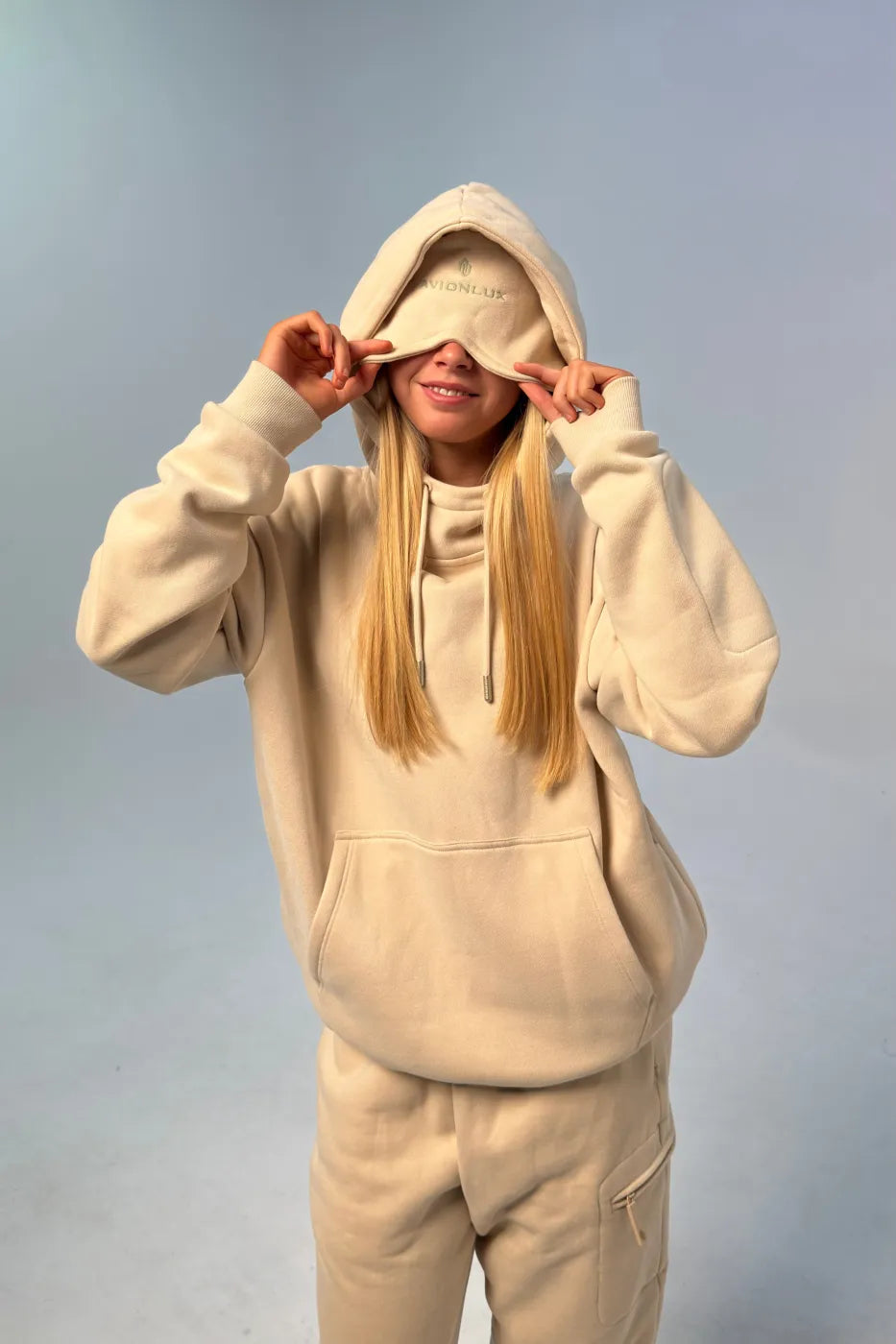 Person wearing a beige hoodie with eye mask and pants with a plain background