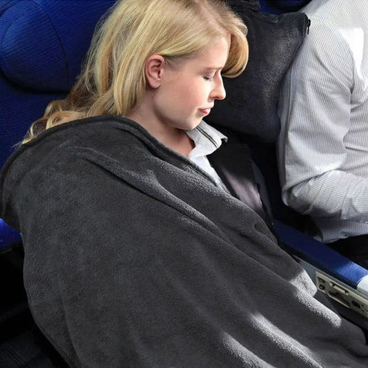 A blonde woman resting her head wrappted in a grey Cabeau Fold And Go Travel Blanket while seated in an airplane cabin next to another passenger.