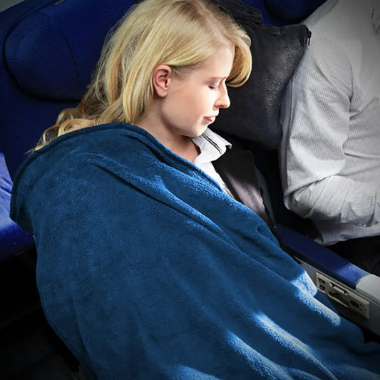 A blonde woman resting on a train or plane, wrapped in a plush blue Cabeau 4 in 1 Fold and Go Travel blanket. She is seated next to another passenger in a white shirt, with blue upholstered seats visible in the background.