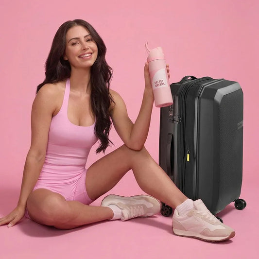 A woman in a pink bodysuit sits on a pink studio floor next to a black Delsey Anvers 55cm suitcase, holding a pink reusable water bottle with the words 'Delsey Anvers.