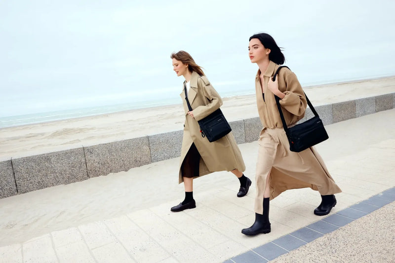 Hedgren EYE NEO Medium Crossbody Bag with RFID Pocket in Green Milieu, worn on the shoulder by two women walking a seafront promenade in trench coats