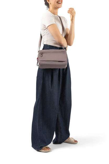 Hedgren EYE NEO Medium Crossbody Bag with RFID Pocket in Willow Grey, worn crossbody at the waist by a woman in a white tee and wide denim trousers