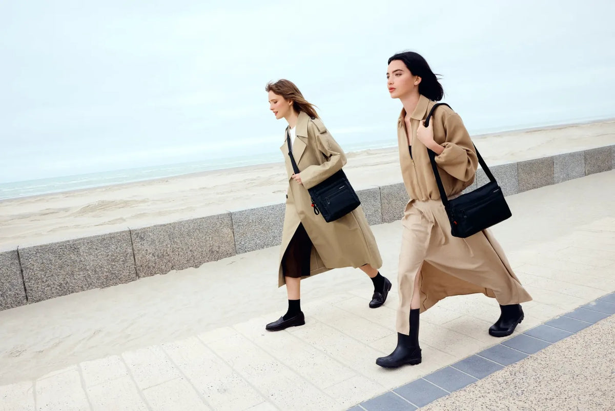 Hedgren EYE NEO Medium Crossbody Bag with RFID Pocket in Willow Grey, worn on the shoulder by two women walking a seafront promenade in trench coats