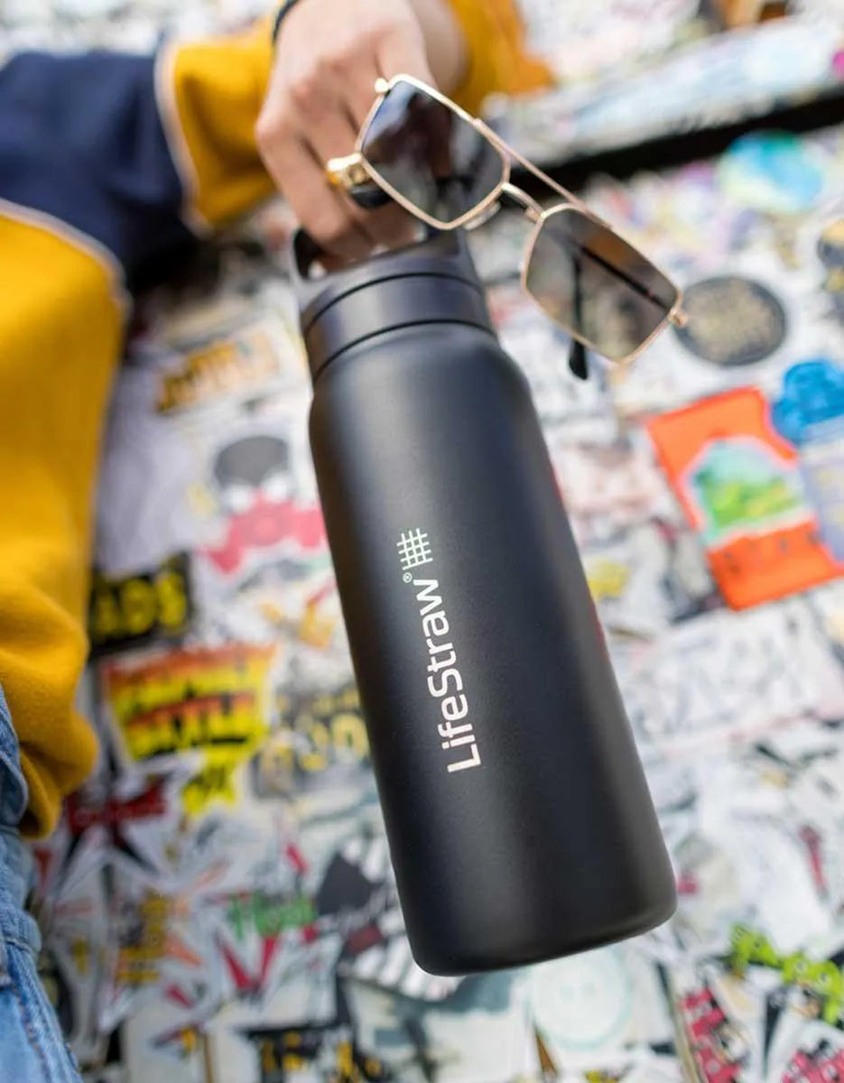 LifeStraw Go 2.0: a black stainless steel LifeStraw Go bottle held by its handle over a sticker-covered wall