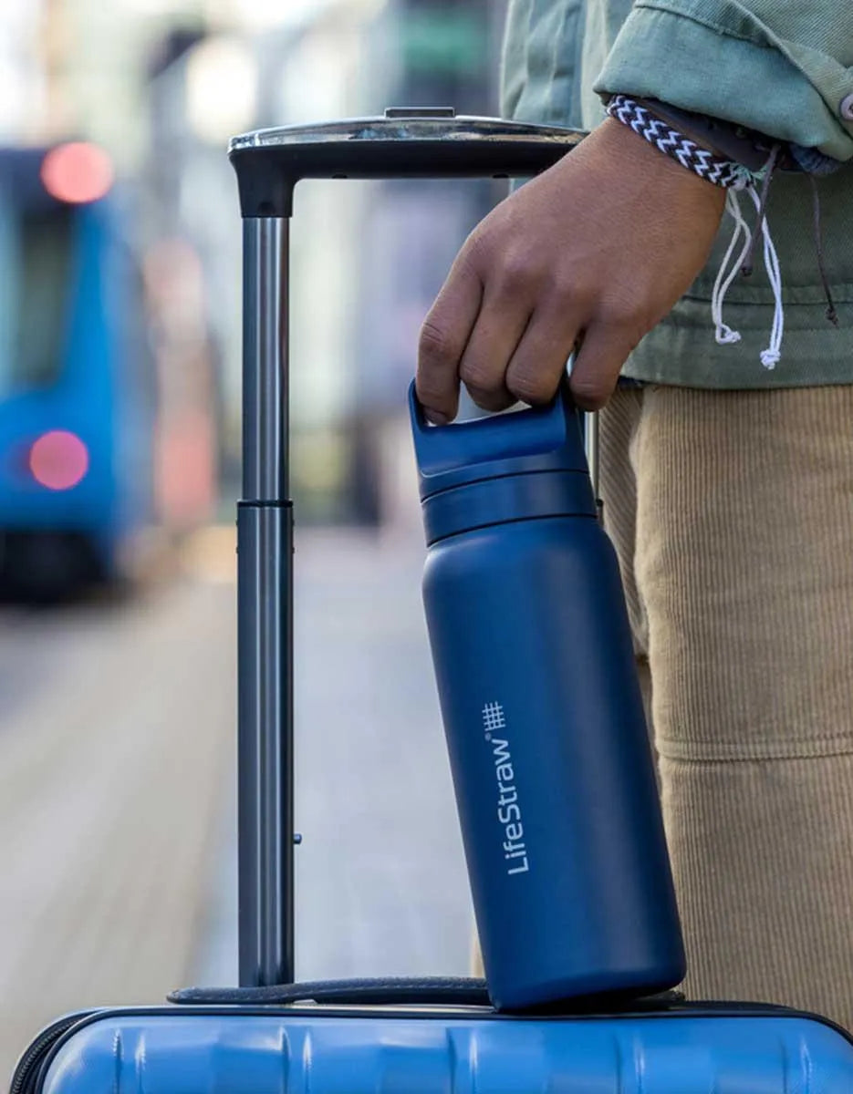 LifeStraw Go 2.0: a navy stainless steel LifeStraw Go bottle resting on a blue suitcase as a traveller holds the handle