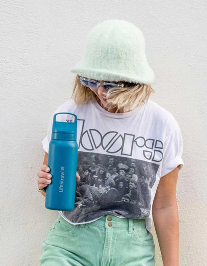 LifeStraw Go 2.0: a stainless steel LifeStraw Go bottle in Laguna Teal held by a woman in a bucket hat and band T-shirt against a white wall