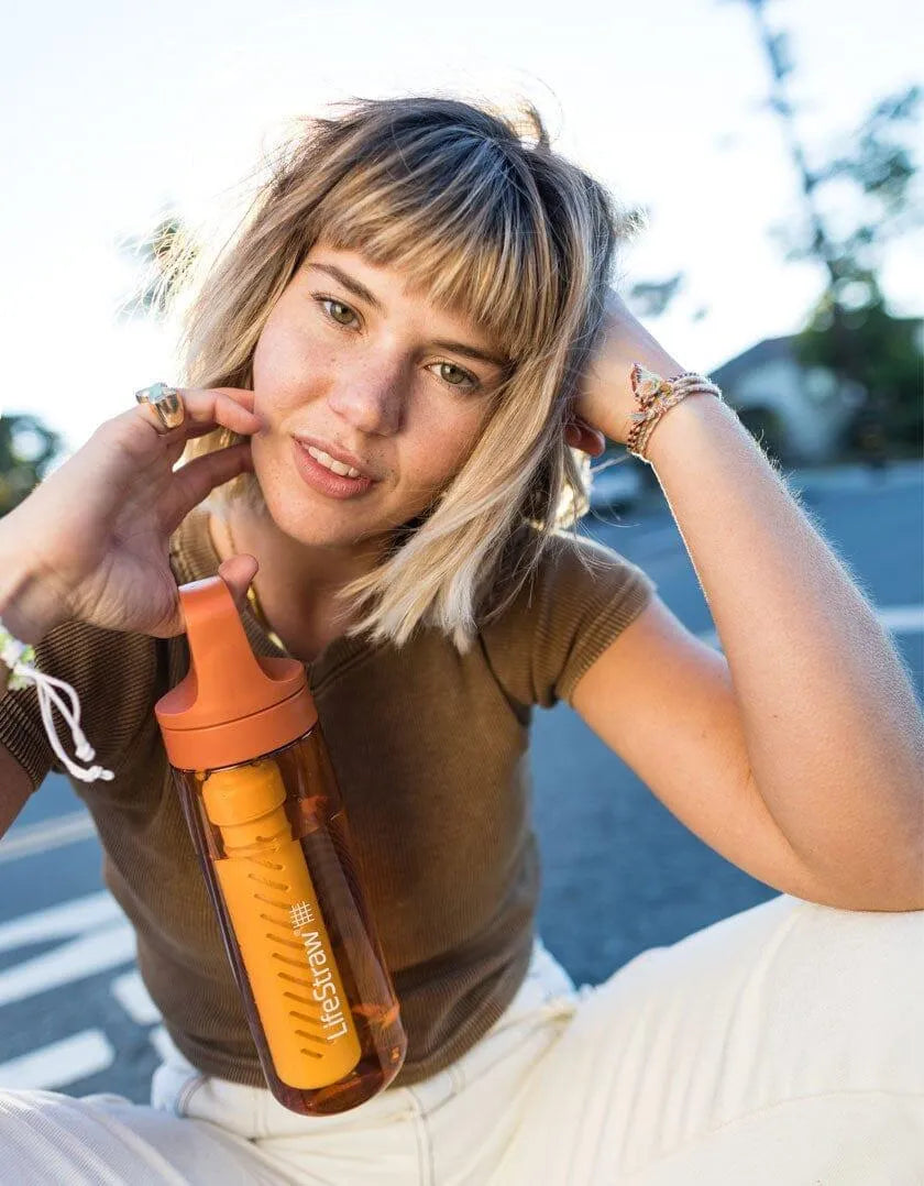 LifeStraw Go 2.0: a smiling woman sitting in the street sun holding an orange LifeStraw Go bottle