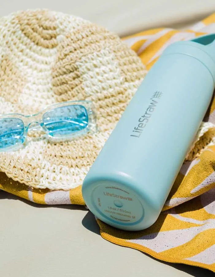 LifeStraw Go 2.0: a pale green LifeStraw Go 500ml stainless steel bottle lying on a straw hat and beach towel beside sunglasses