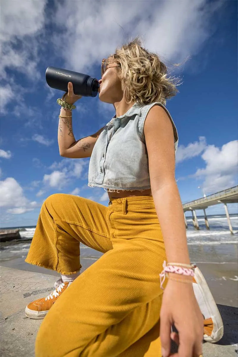 LifeStraw Go 2.0: a woman drinking from a black stainless steel LifeStraw Go bottle on a sunny beach