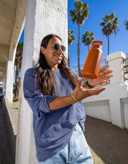 LifeStraw Go 2.0: a woman tossing an orange stainless steel LifeStraw Go bottle beside a palm-lined seaside walkway