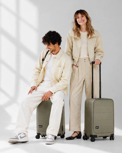 Couple relaxing, one seated on the MiaMily Carry-on Seat Pro suitcase in Olive, showing the patented built-in seat