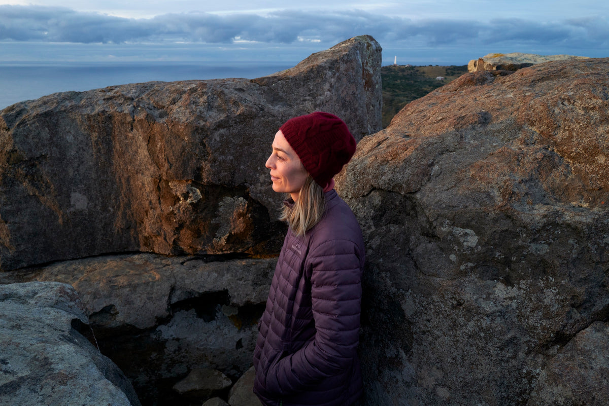 Walker in a beanie beside lichen-covered boulders above the sea at dusk