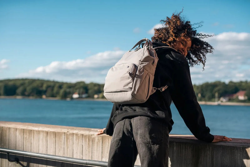 Pacsafe Citysafe CX Anti-Theft 16" Laptop Backpack in Gravity Grey, worn on both shoulders by a woman leaning on a waterfront railing