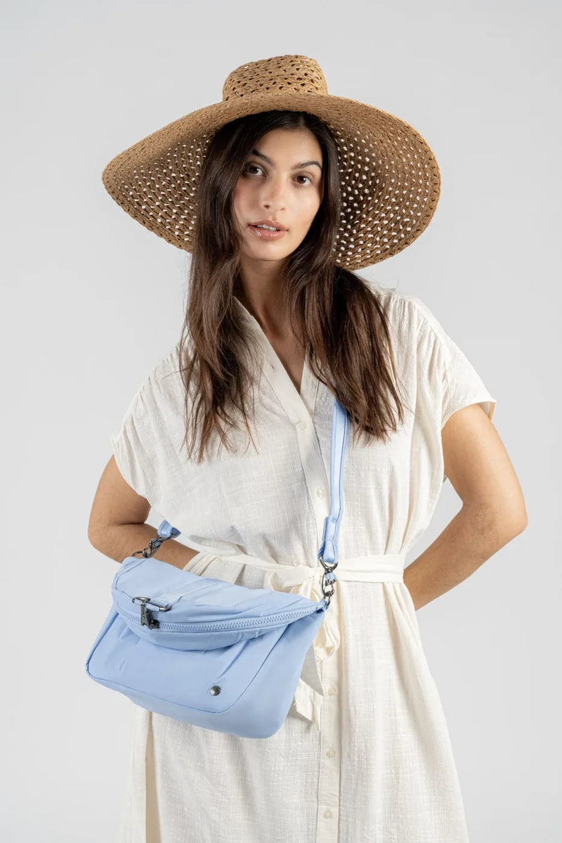 Pacsafe Citysafe CX Anti-Theft Convertible Backpack / Shoulder Bag in Serenity Blue, worn crossbody at the hip by a woman in a cream dress and straw hat