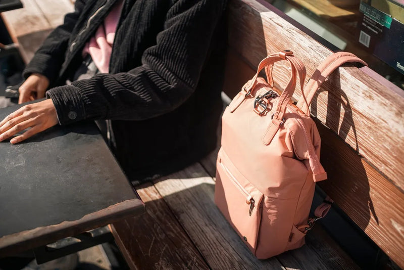 Pacsafe Citysafe CX Econyl Anti-Theft Mini Backpack in Rose, set down on a timber bench beside a seated woman