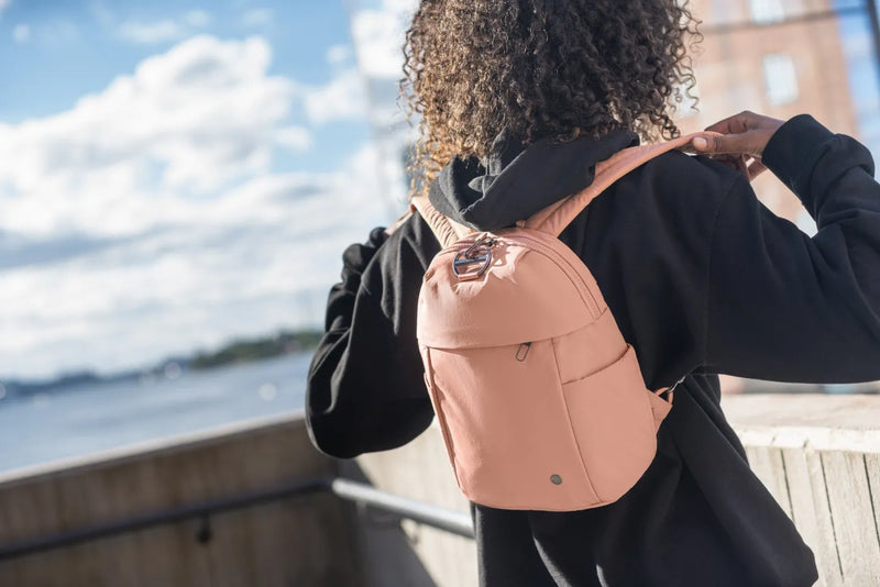 Pacsafe Citysafe CX Petite Anti-Theft 8L Backpack in Rose, worn on both shoulders by a woman on a waterfront walkway
