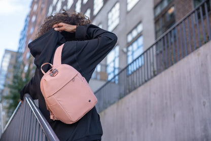 Pacsafe Citysafe CX Petite Anti-Theft 8L Backpack in Rose, swung off one shoulder by a woman on a city street