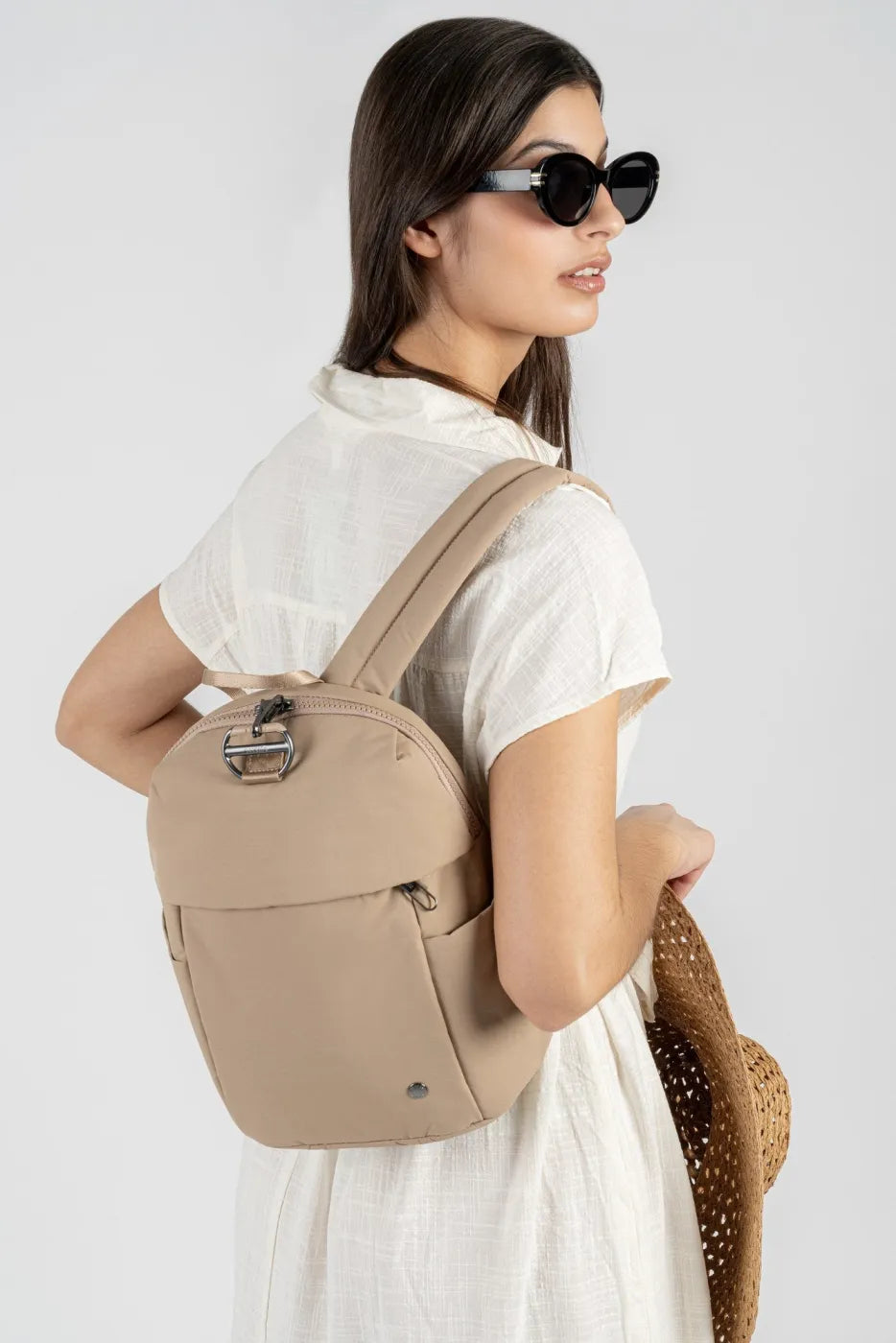 Pacsafe Citysafe CX Petite Anti-Theft 8L Backpack in Taupe, worn on both shoulders by a woman in a white dress carrying a straw hat
