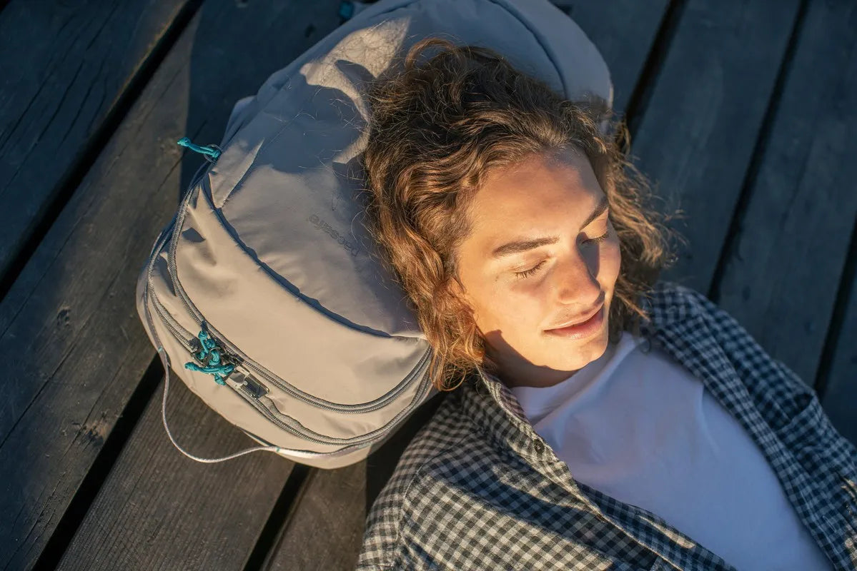 Pacsafe Eco 25L Anti-Theft 16" Laptop Backpack in Gravity Grey, used as a pillow by a woman lying on a sunlit jetty