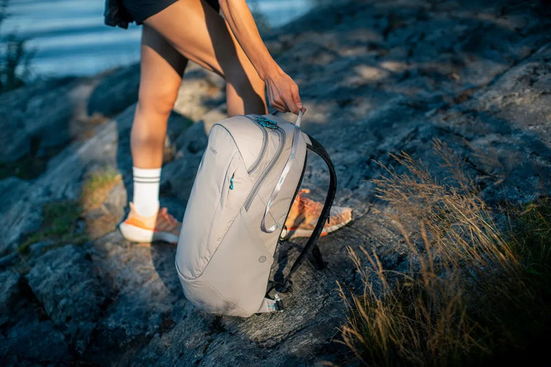 Pacsafe Eco 25L Anti-Theft 16" Laptop Backpack in Gravity Grey, standing on a rock by the water while its owner steps past