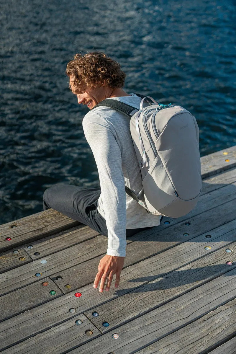 Pacsafe Eco 25L Anti-Theft 16" Laptop Backpack in Gravity Grey, worn on both shoulders by a man sitting on a boardwalk by the sea