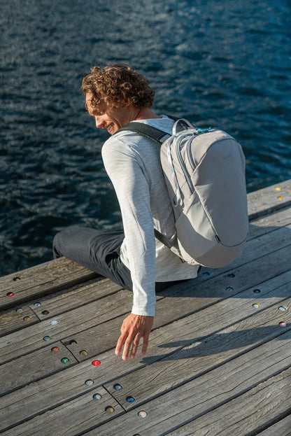 Pacsafe Eco 25L Anti-Theft 16" Laptop Backpack in Gravity Grey, worn on both shoulders by a man sitting on a boardwalk by the sea