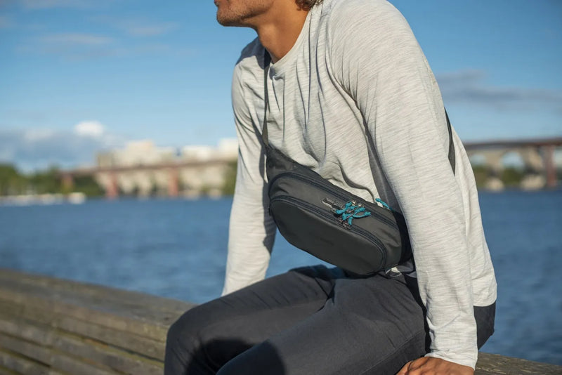 Pacsafe ECO Anti-theft Waist Pack in Black, worn crossbody by a man sitting on a jetty by the water