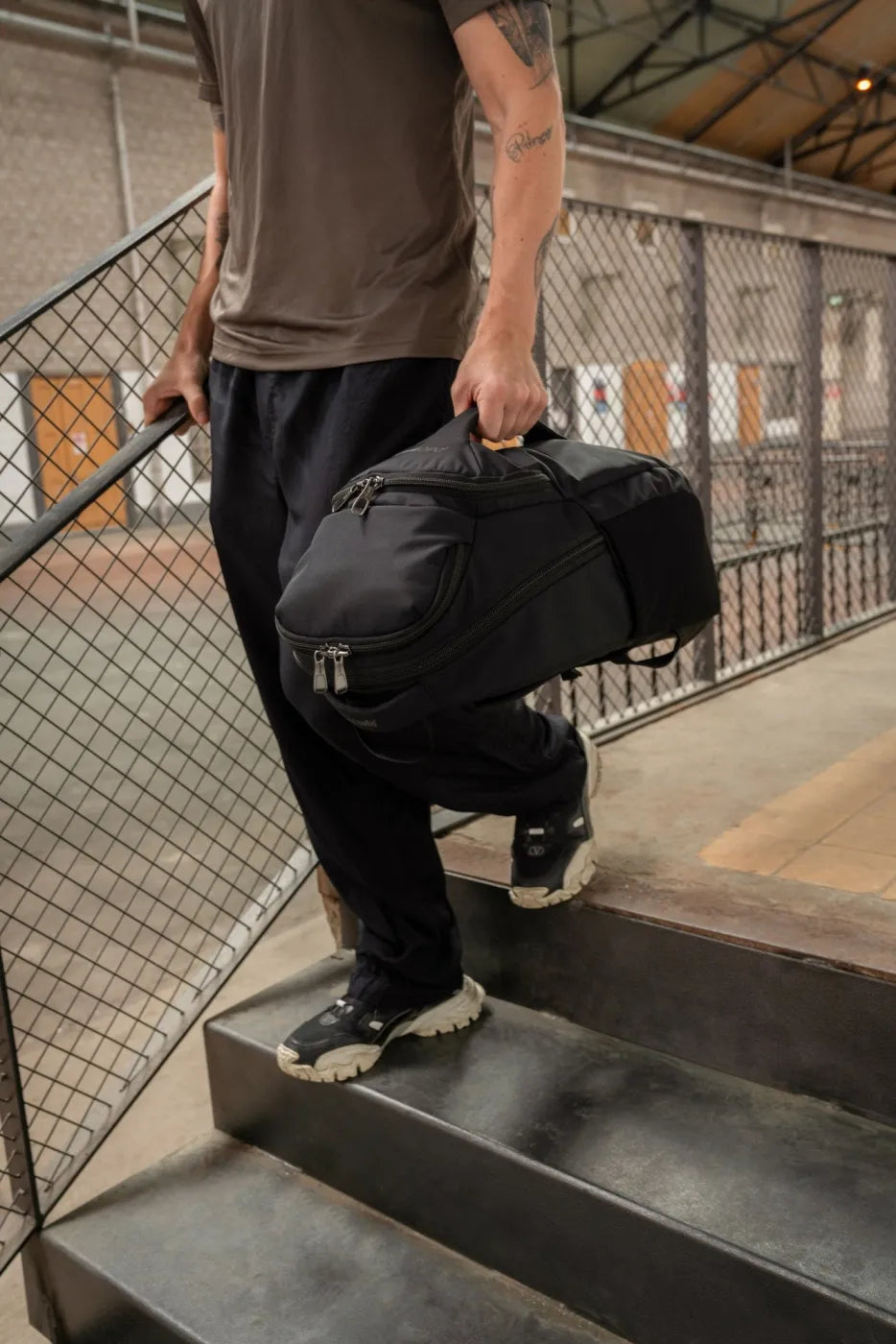 Pacsafe EXP Anti-Theft 28L 16" Laptop Backpack in Black, carried by the grab handle by a man walking down steps in a station