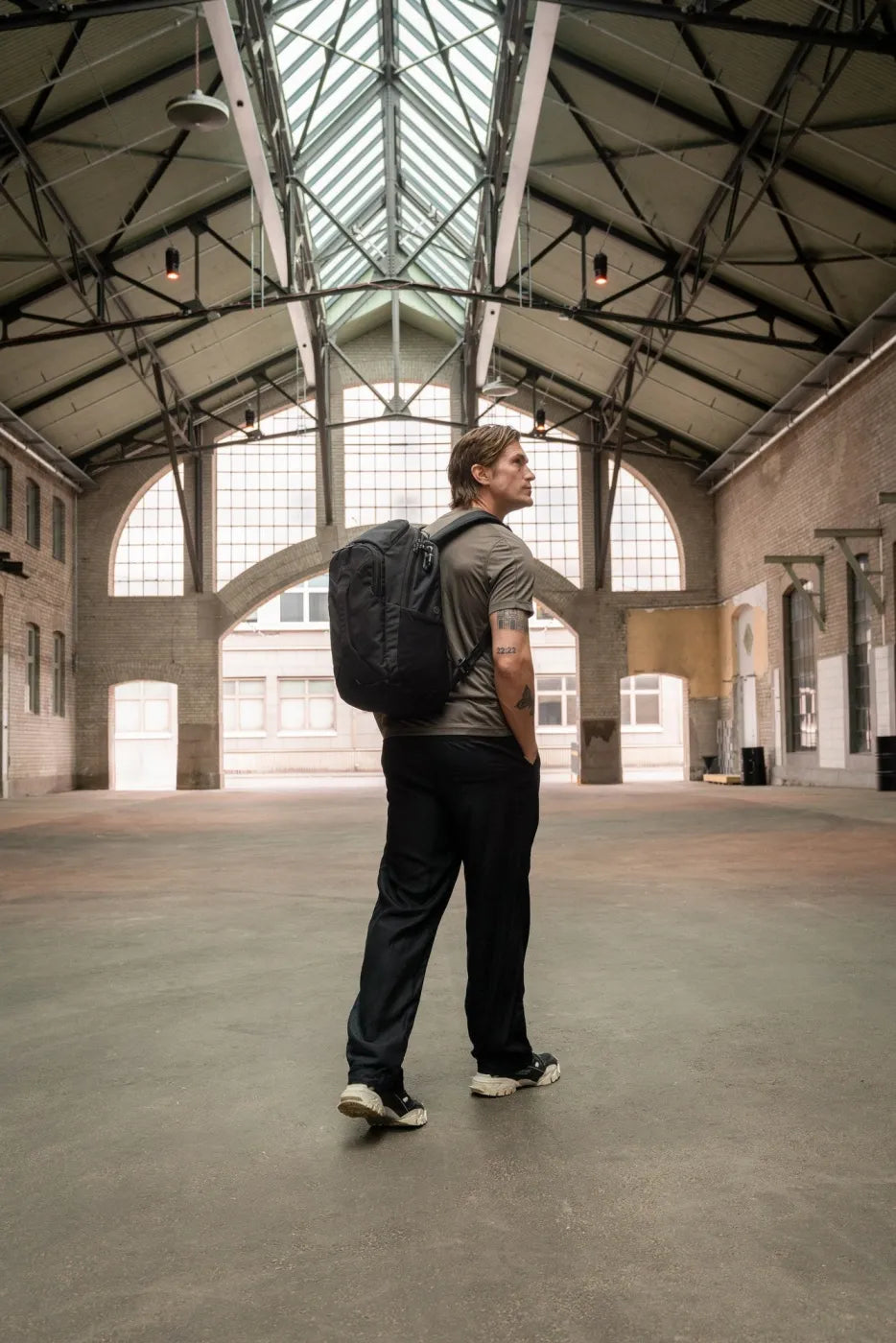 Pacsafe EXP Anti-Theft 28L 16" Laptop Backpack in Black, worn on both shoulders by a man crossing an old industrial hall