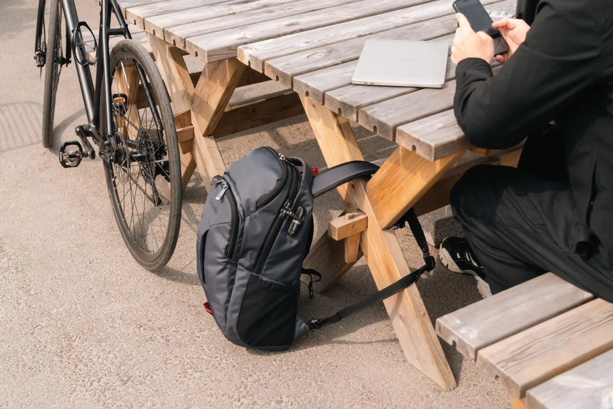 Pacsafe EXP Anti-Theft 28L 16" Laptop Backpack in Slate, propped against a picnic table beside a bicycle while its owner works