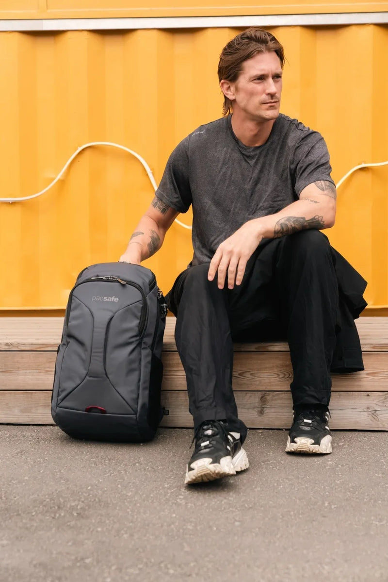 Pacsafe EXP Anti-Theft 28L 16" Laptop Backpack in Slate, standing on the ground beside a man sitting against a yellow shipping container