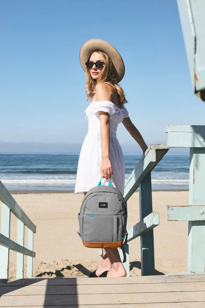 Pacsafe GO 15L Anti-Theft 13" Laptop Backpack in Stone, carried by the grab handle by a woman on beach steps