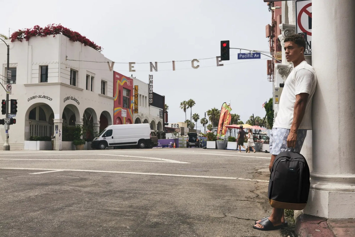 Pacsafe GO 25L Anti-Theft 16" Laptop Backpack in Jet Black, held at the side by a man leaning against a pillar in Venice Beach