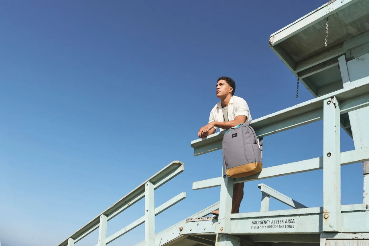 Pacsafe GO 25L Anti-Theft 16" Laptop Backpack in Stone, resting on a lifeguard tower railing beside a man looking out to sea