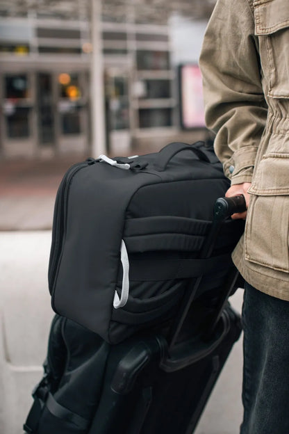 Pacsafe GO 34L Anti-Theft Carry-on Backpack in Jet Black, hooked over the handle of a wheeled case at an airport kerb