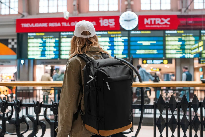 Pacsafe GO 44L Anti-Theft Carry-on Backpack in Jet Black, worn on both shoulders by a woman reading a station departure board