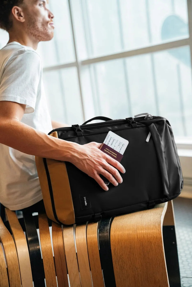 Pacsafe GO 44L Anti-Theft Carry-on Backpack in Jet Black, held on the lap of a man in an airport lounge, boarding pass in hand