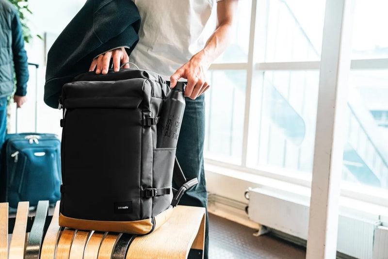 Pacsafe GO 44L Anti-Theft Carry-on Backpack in Jet Black, set on a bench beside a case at a ferry terminal window
