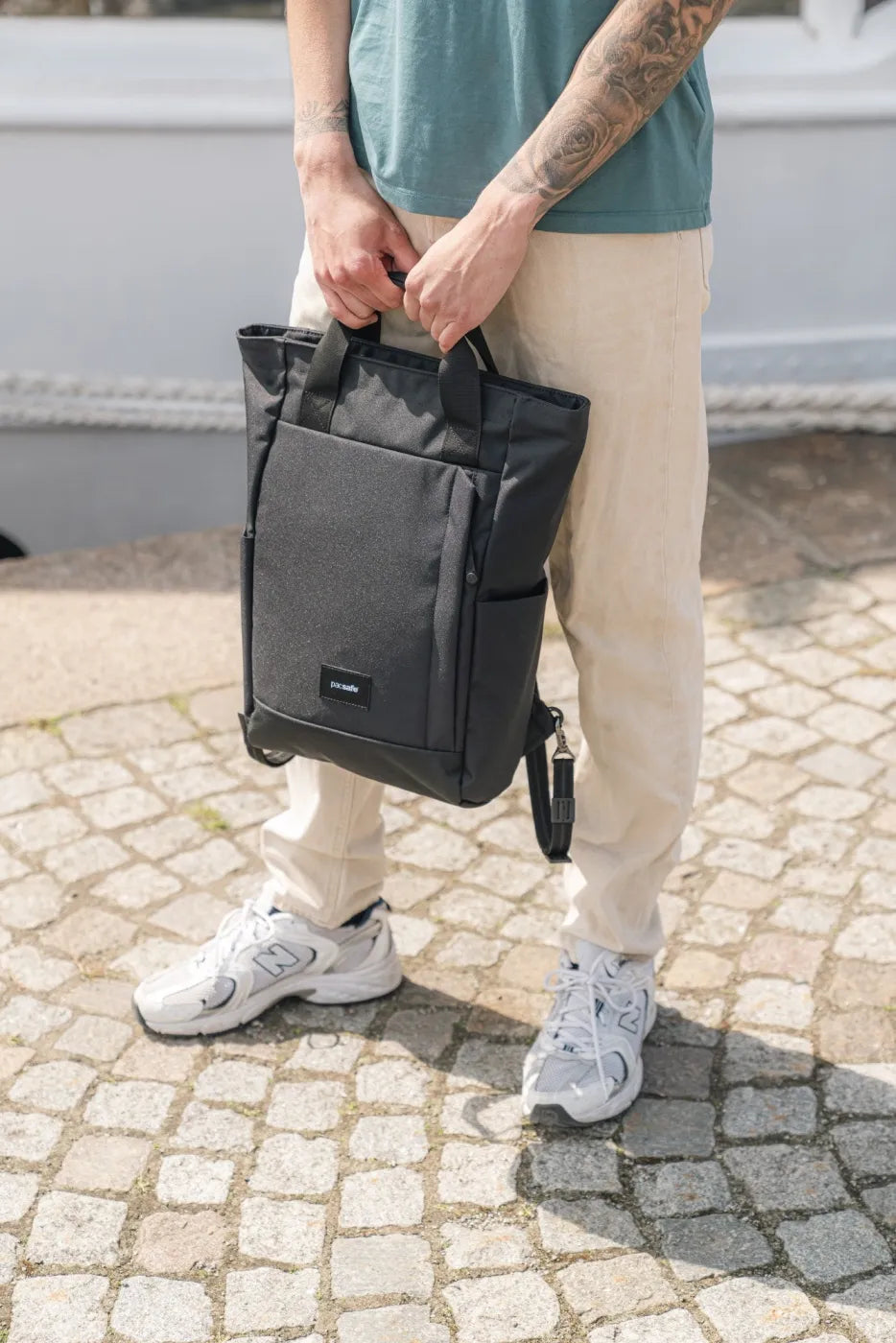 Pacsafe GO Anti-Theft 16" Laptop Totepack in Jet Black, held by both top handles in front of a man on a cobbled street