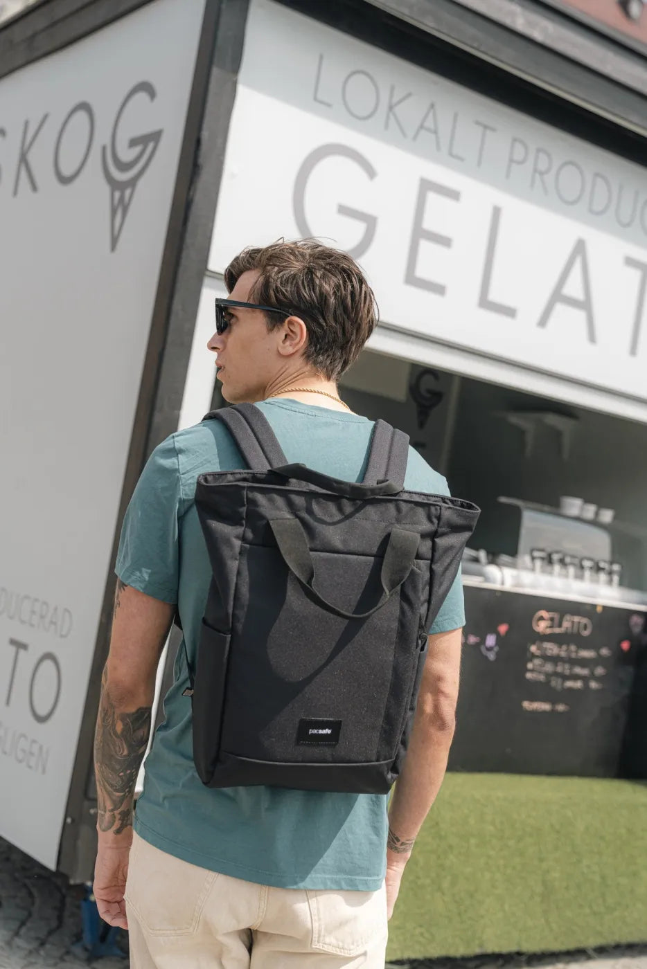 Pacsafe GO Anti-Theft 16" Laptop Totepack in Jet Black, worn as a backpack by a man standing at a gelato kiosk