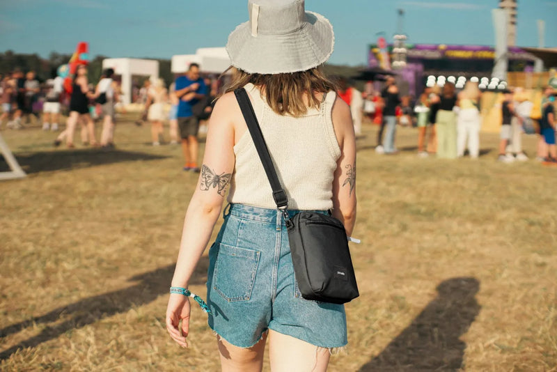Pacsafe Go Anti-theft Festival Crossbody Bag in Jet Black, worn crossbody at the hip by a woman walking through a festival field
