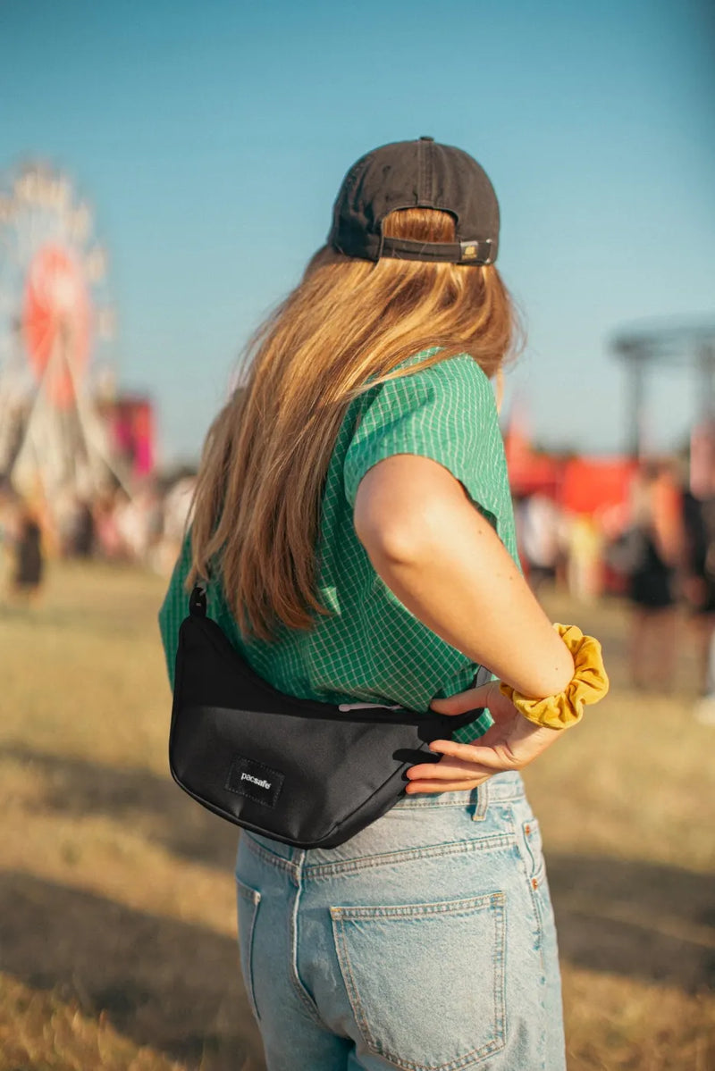Pacsafe Go Anti-Theft Lunar Crossbody Bag in Jet Black, worn at the back of the waist by a woman at a festival, seen from behind
