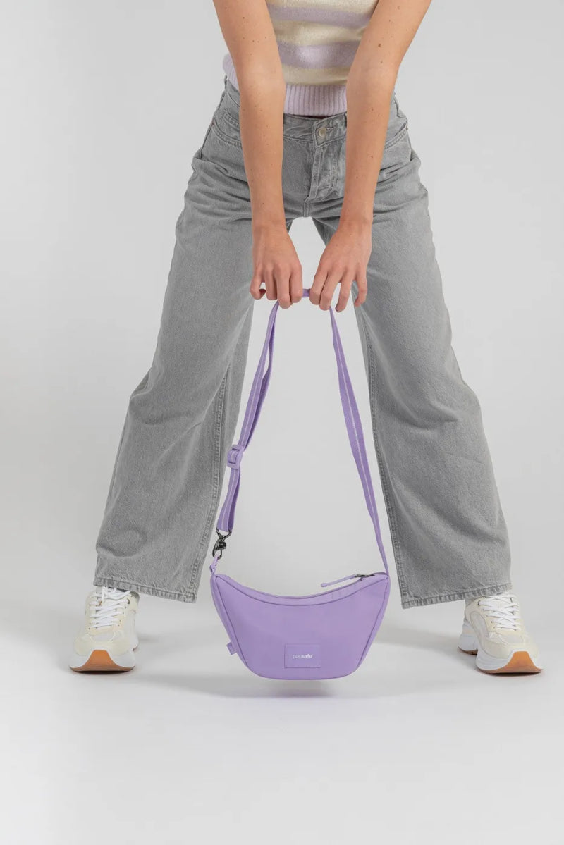 Pacsafe Go Anti-Theft Lunar Crossbody Bag in Lavender, hanging from its strap at knee height, held by a woman in grey wide leg jeans