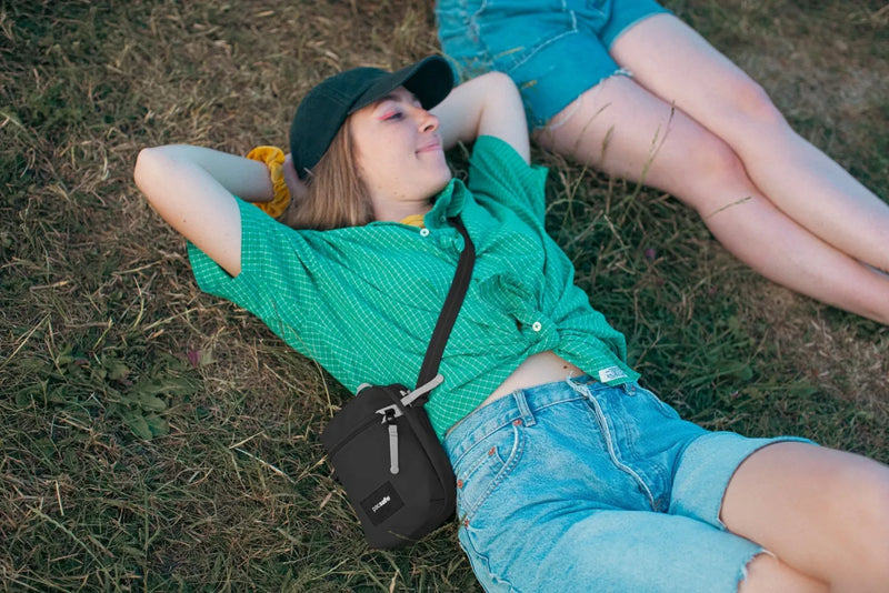 Pacsafe Go Anti-theft Micro Crossbody Bag in Jet Black, worn crossbody by a woman lying back on the grass at a festival