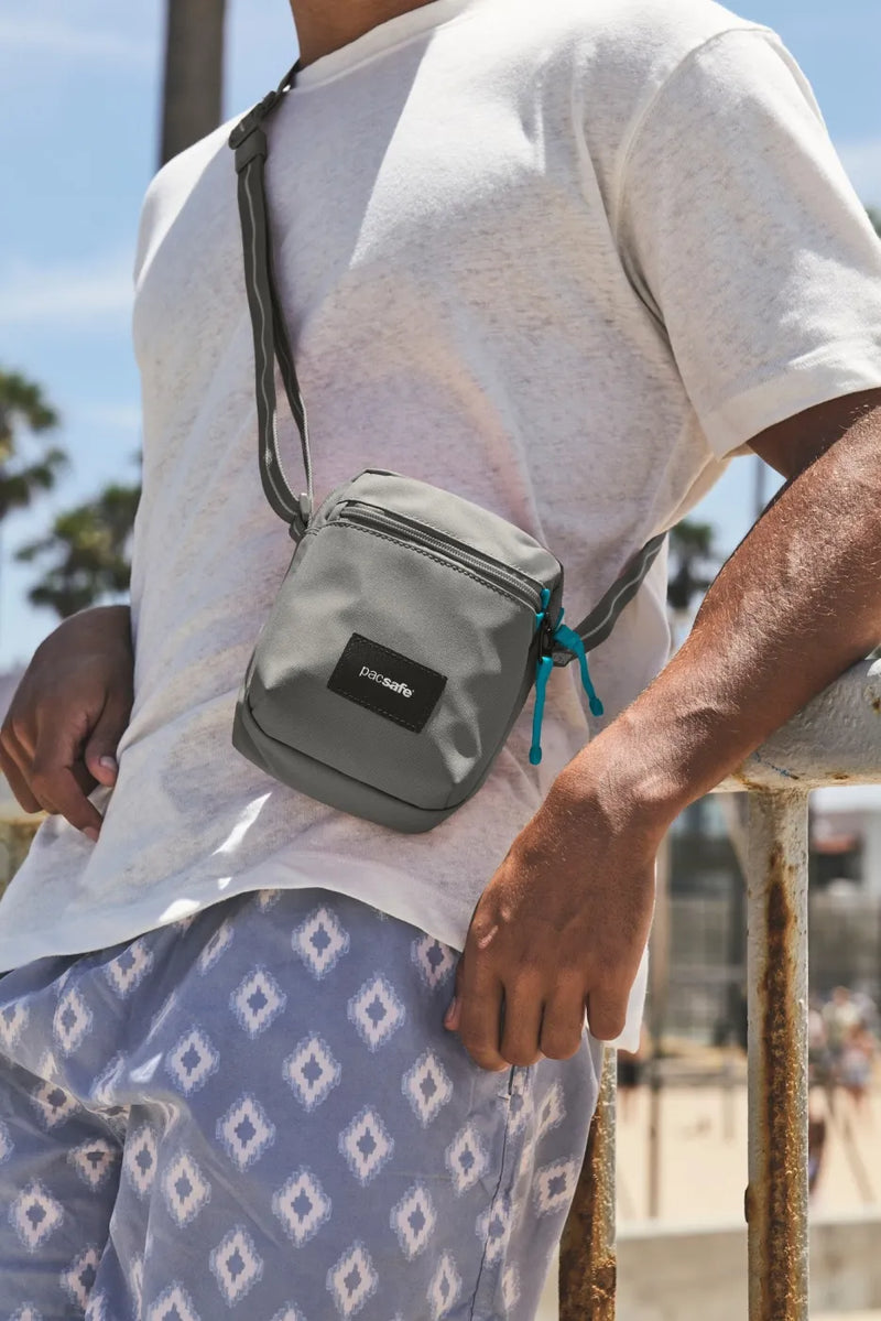 Pacsafe Go Anti-theft Micro Crossbody Bag in Stone, worn crossbody by a man leaning on a beachfront railing