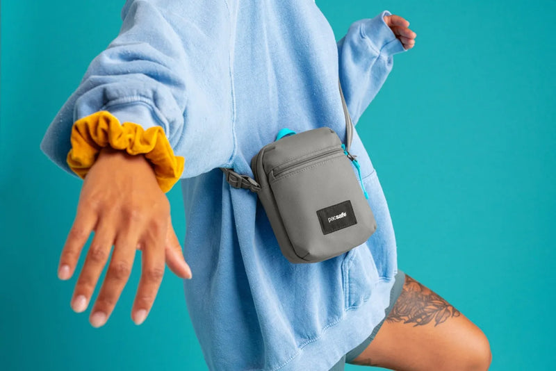 Pacsafe Go Anti-theft Micro Crossbody Bag in Stone, worn crossbody at the hip over a blue shirt, against a teal backdrop
