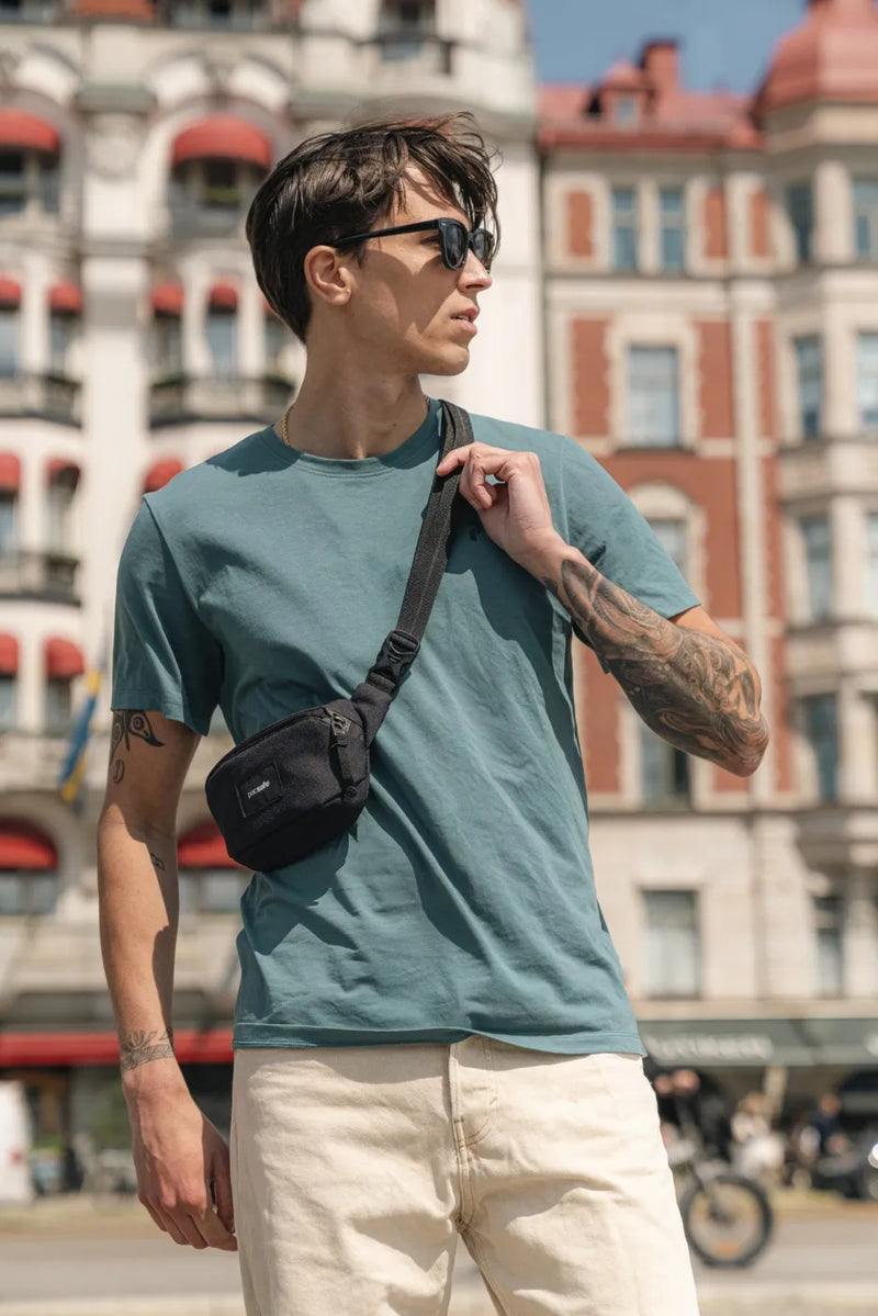 Pacsafe Go Anti-Theft Micro Sling in Jet Black, worn crossbody by a man adjusting the strap on a city street