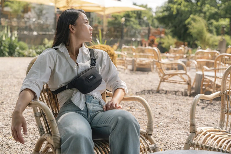 Pacsafe Go Anti-Theft Micro Sling in Jet Black, worn across the chest by a woman sitting in a cane chair at an outdoor bar