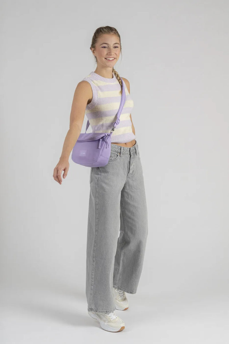 Pacsafe Go Anti-Theft Saddle Crossbody Bag in Lavender, worn crossbody at the hip by a woman walking in grey jeans