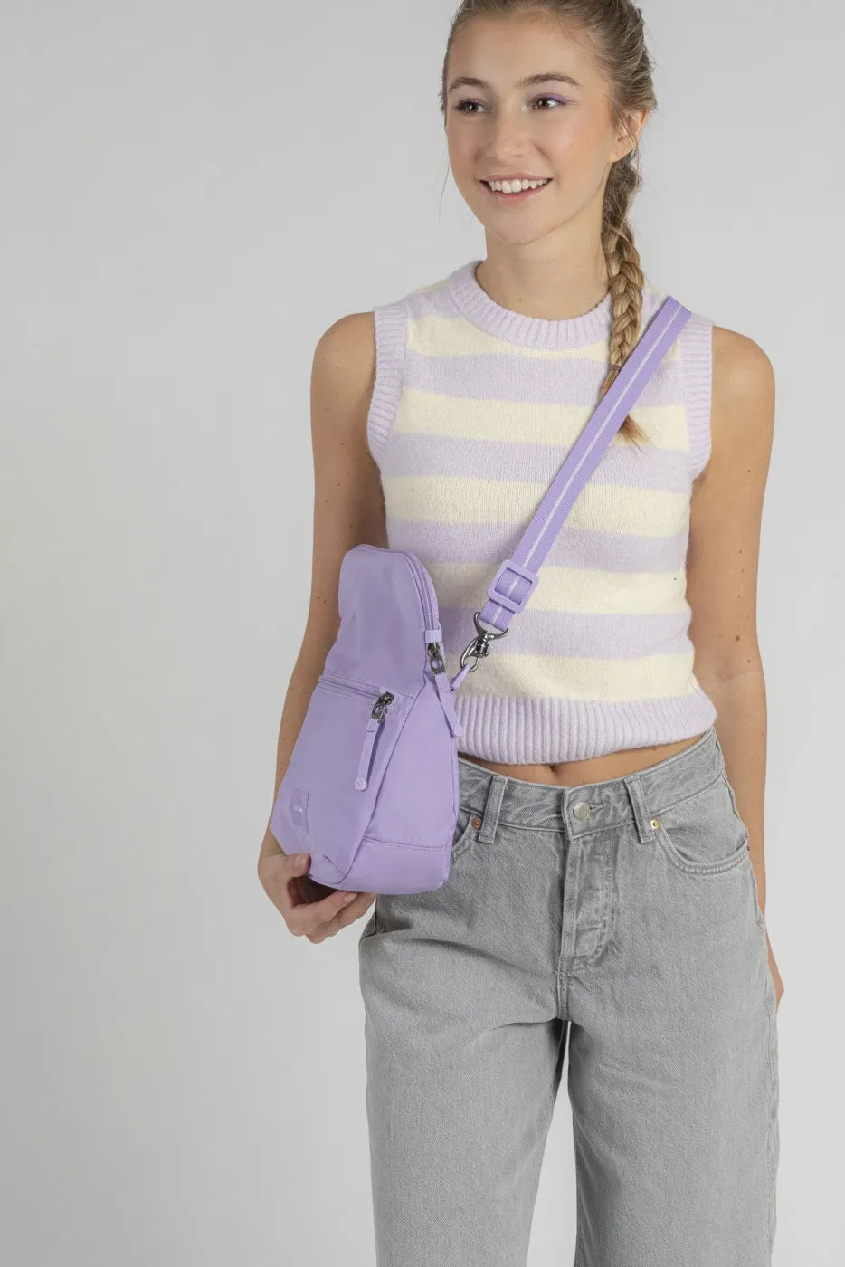 Pacsafe Go Anti-Theft Saddle Crossbody Bag in Lavender, held at the hip by a woman in a striped knit vest and grey jeans